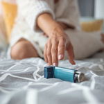 A person wearing white, sitting on a bed, reaching a hand towards a blue inhaler.