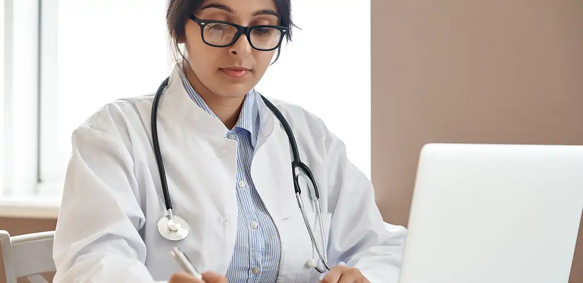 A doctor sat in front of a laptop, writing on a notepad.