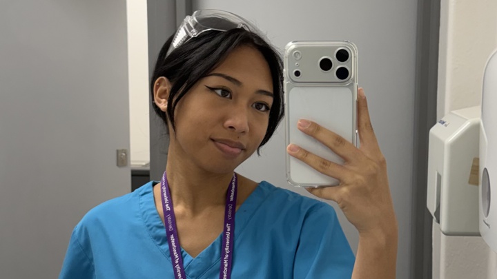 Student Ashleigh in blue dental scrubs holding her phone up to take a photo in a mirror.