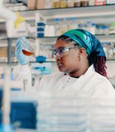 A female student in the lab wearing a white lab coat looks into a conical flask.