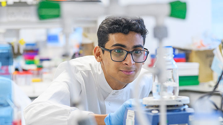A student in a lab coat and glasses is looking across a lab table.