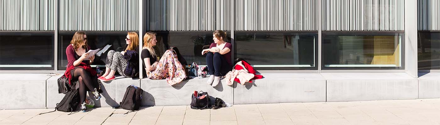 A group of young people sat outdoors against a building talking and studying in the sunshine.