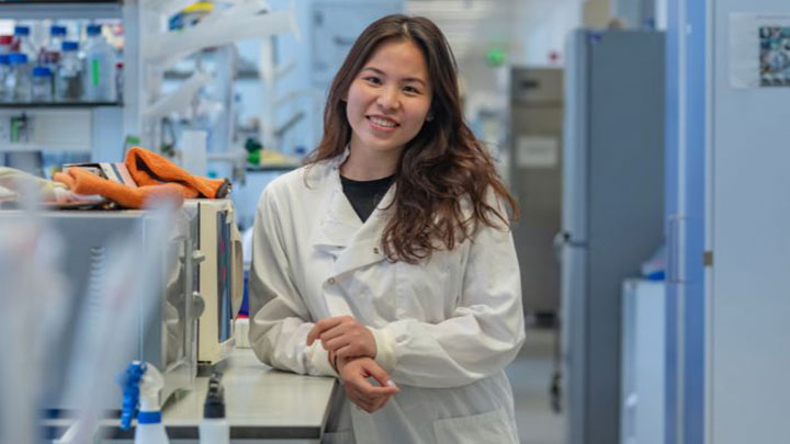 A postgraduate researcher standing in a lab, smiling at the camera.