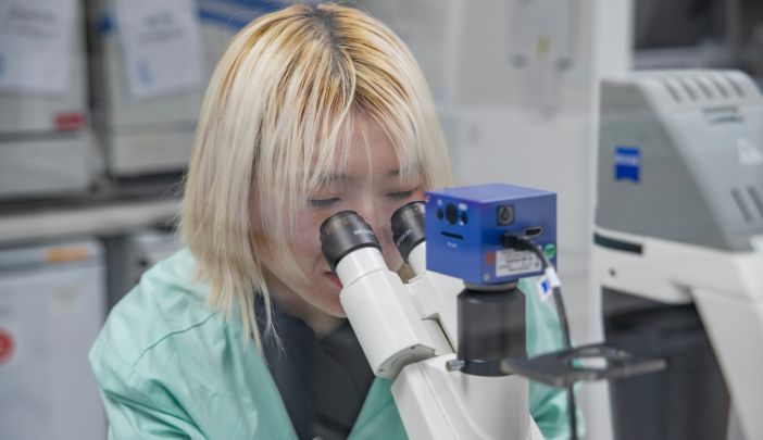 A postgraduate researcher in a lab coat looking through a microscope.