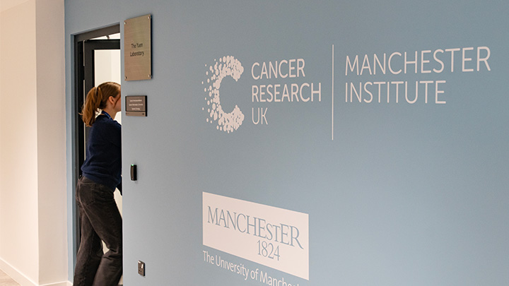 Researcher walking through doorway with university and CRUK MI logos on wall