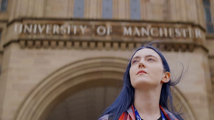 Portrait of a woman standing outside of a building with The University of Manchester written on it. 