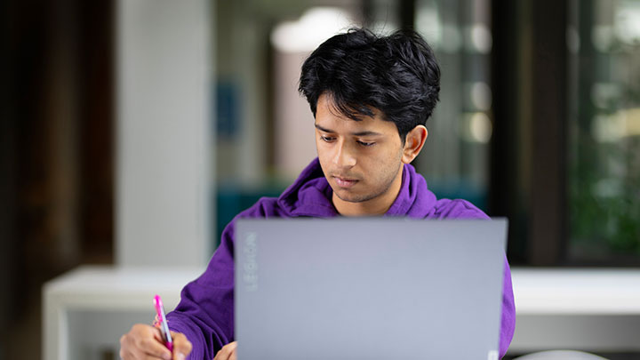 A student wearing a purple jumper working on a laptop and writing in a notebook.