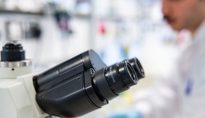 A microscope in the foreground with a postgraduate researcher in the lab in the backgoround