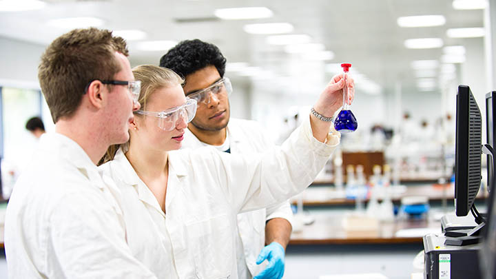 Three researchers in lab coats looking at a flask with blue liquid.