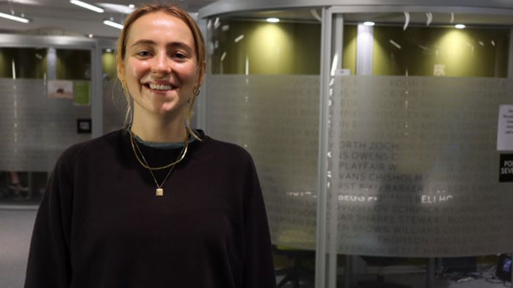 Postgraduate researcher Katie smiling at the camera standing in a slightly dark office space.