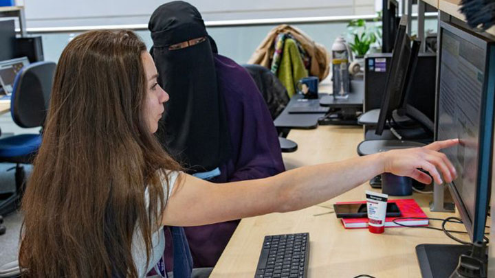 Two students working together at a computer screen. One of the students is pointing at the screen.