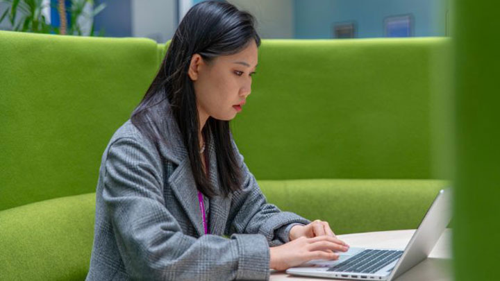 A postgraduate researcher sitting on a green chair working on a laptop.