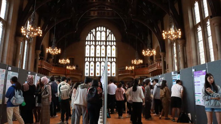 Students are looking at boards and posters placed within an old hall with stained glass windows.